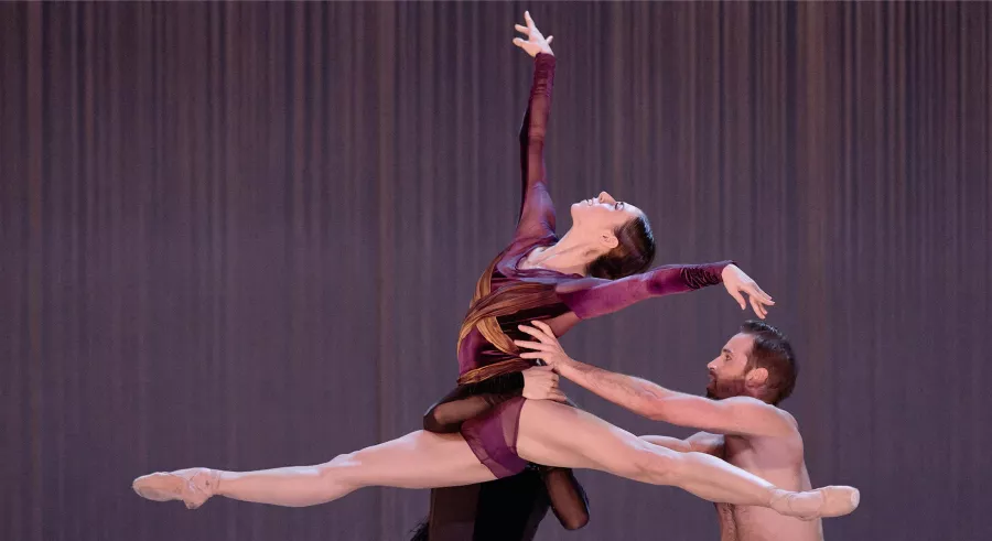 Three dancers appear on stage wearing black, gold, and purple clothing. One dancer another in a lift, with the third dancer out of view behind them.