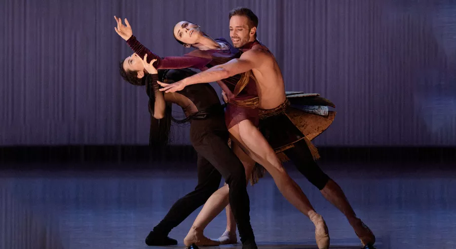 Three dancers appear on stage wearing black, gold, and purple clothing. One dancer leans backwards with the other two leaning against them towards the left of the frame.