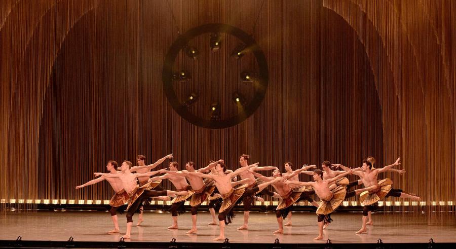 A group of dancers appear onstage in rows of horizontal lines with one leg planted on the ground and the other lifted parallel to the stage. Their arms reach up and out to the sides.