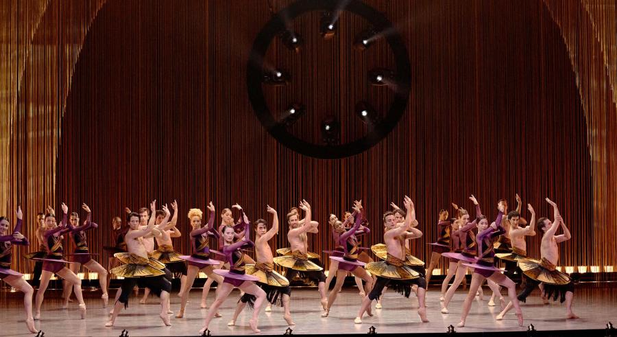 A group of dancers dressed in purple and gold and standing in multiple rows horizontal lines stand with their feet planted on the ground with a slight bend at one knee. They reach their arms up, with one placed at the elbow of the other.
