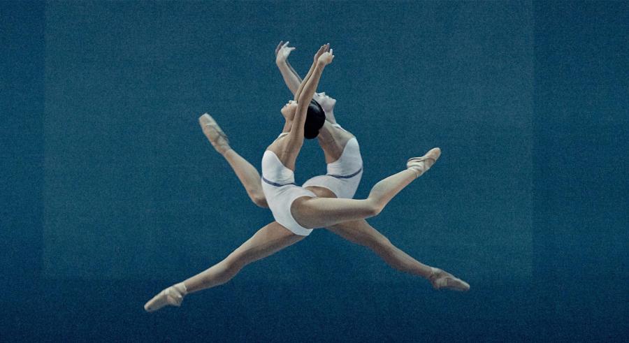 Two dancers dressed in white leap in-air across the stage, appearing directly centered and facing opposite directions.