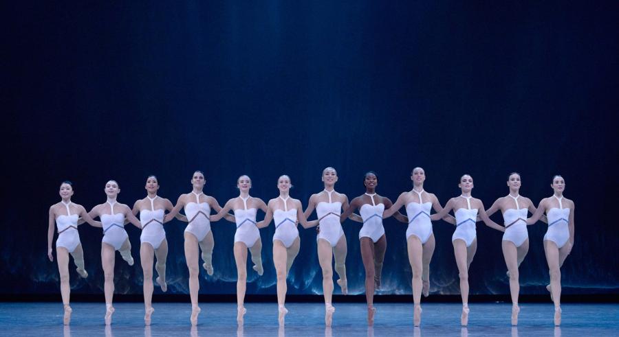 A group of dancers dressed in matching white clothing stand on a blue-lit stage in a line with their arms linked and one leg lifted up behind them.