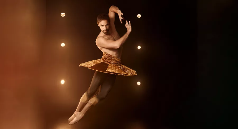 A dancer dressed in a structured tutu-like piece with a bare chest twirls in the air onstage with a glow of soft yellow lighting behind them.