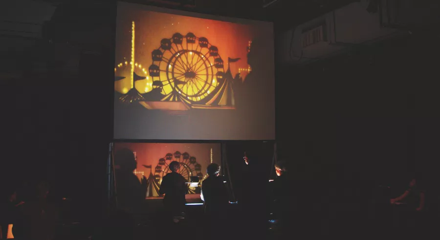 A projected screen shows a ferris wheel and tents with an orange-yellow background. Performers below act live in front of a film production station, which appears on-screen.