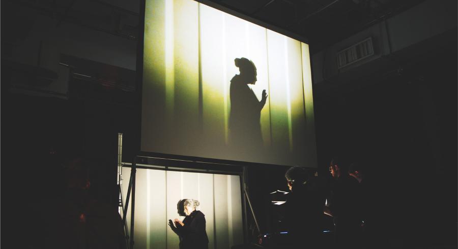 A performer stands in front of a film production station and appears projected above on a screen, displaying them as a shadowed figure against bright yellow lighting.