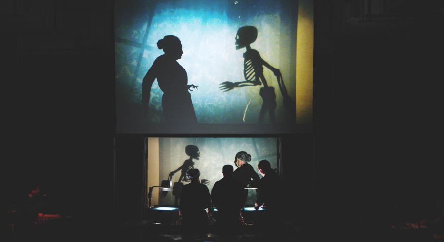 One performer and a skeleton figure stand facing one another in front of a film production station and appear projected above on a screen, displaying them as shadowed figures against blue lighting.