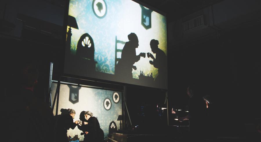Two performers stand facing one another in front of a film production station and appear projected above on a screen, displaying them as shadowed figures within a living room setting. 