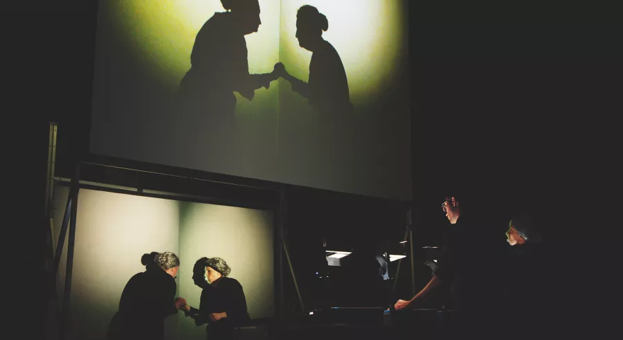 Two performers stand in front of a film production station and appear projected above on a screen that displays them as shadowed figures looking and reaching toward one another with a bright yellow circle behind.