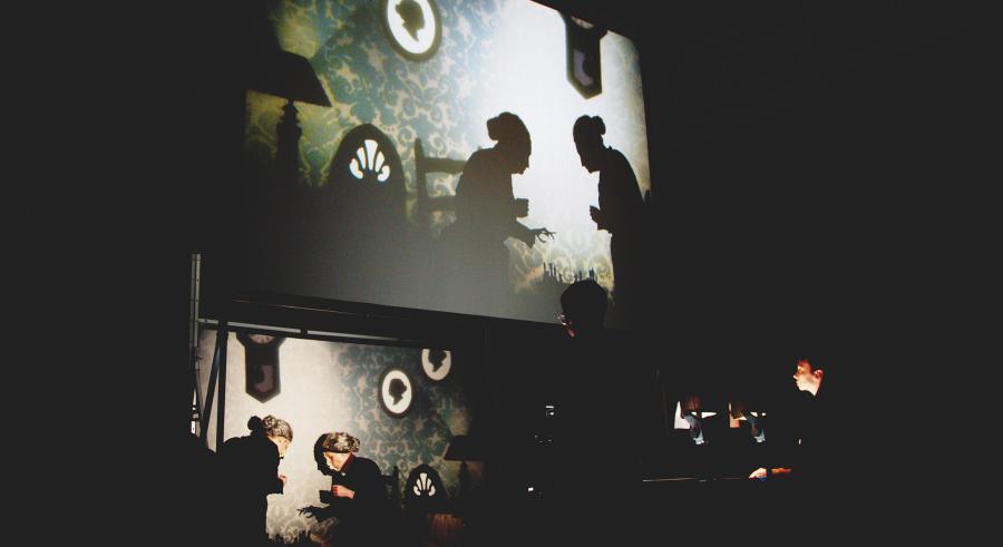 Two performers stand in front of a film production station and appear projected above on a screen, displaying them as shadowed figures within a living room setting. 