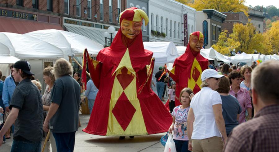 Two very tall puppet figures dressed as jesters in red and yellow robes walk through a busy festival.