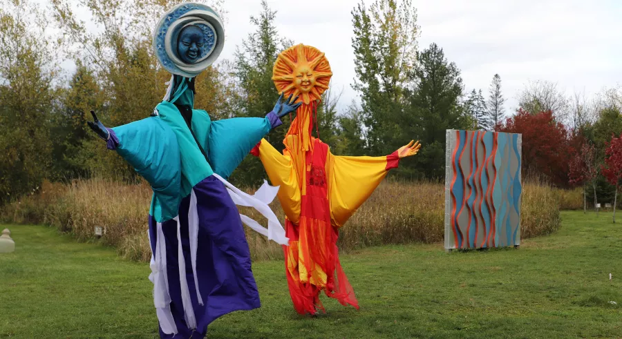 Two very tall colorful puppet figures with images of the moon and sun for faces stand with arms outstretched.