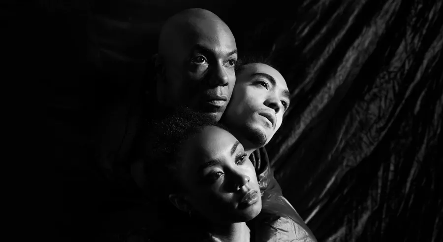 Three people's heads appear against a dark curtain, two direcly placed vertically in a line and the other grouped within and to the right of them. Their faces are covered partially by a dark shadow and they look off into the distance.