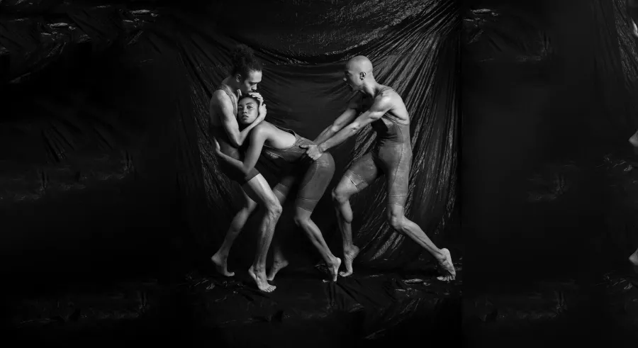 A black and white image shows three dancers in front of a curtain. Two dancers embrace while the other follows closely behind with their hands placed on one of the dancer's back.