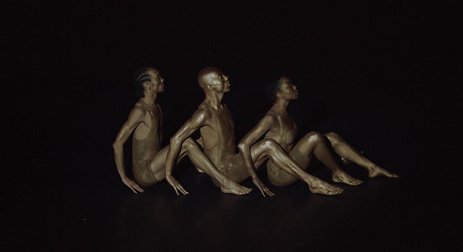 Three dancers appear in dark lighting facing the right and sitting in a line with their legs at the side of one another's bodies. Their hands touch the ground and they look up.