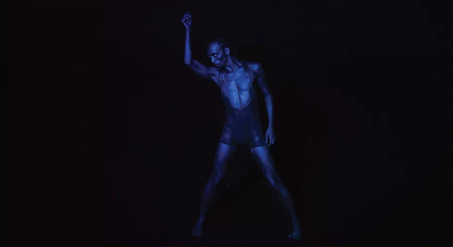 A dancer lit in dark blue lighting stands looking down with their feet planted and one arm lifted up and the other at their side.