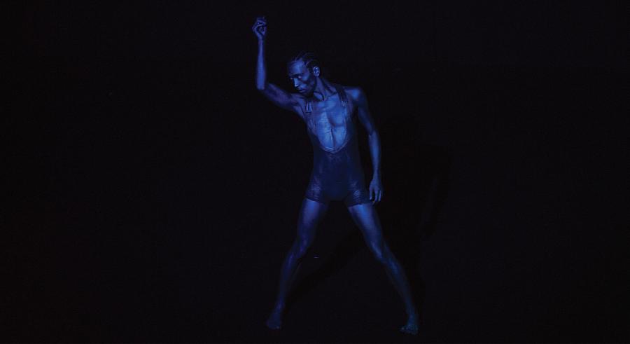 A dancer lit in dark blue lighting stands looking down with their feet planted and one arm lifted up and the other at their side.