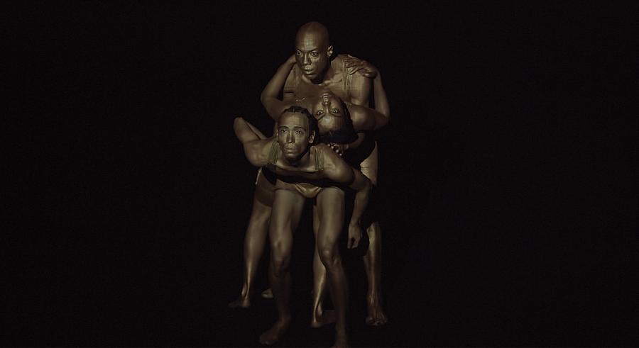 Three dancers appear in dark lighting, facing the camera. One dancer holds another upside down on their back, squished between the thrid dancer who hovers over them.