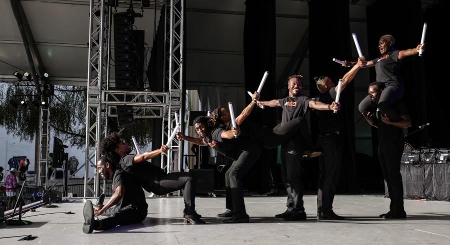 A group of dancers dressed in black form a connected line in various positions, creating a short to tall effect. They hold a sharp object facing upward in their hands.