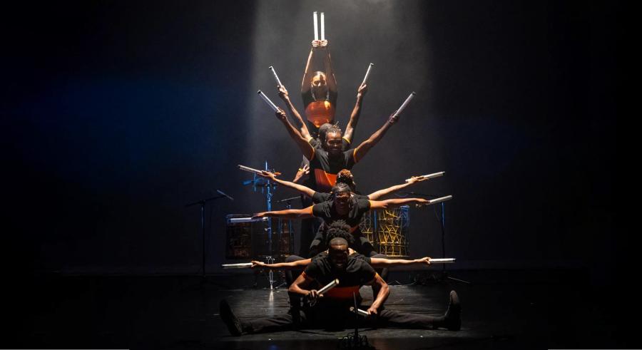 A group of dancers wearing black and red appears onstage vertically in line at different heights, each holding a sharp object pointing outward, forming a semi-circle shape.