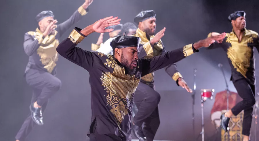 A group of dancers wearing black and gold clothing and caps appears onstage, jumping toward the right of the frame with one hand raised and bent behind them and the other pointed out.