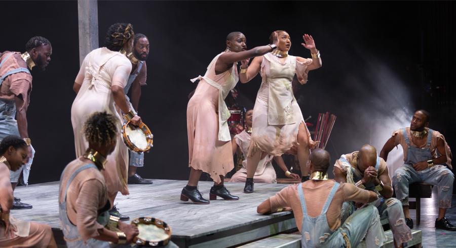 A group of performers appears on stage in various positions wearing a mix of overalls, aprons, and dresses. Two performers appear in the center while the others gather around them, some holding tambourines. 