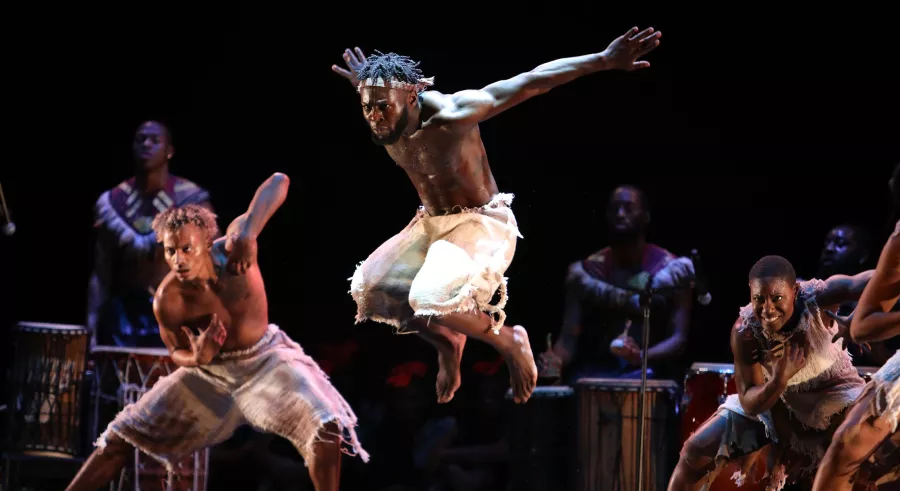 Three dancers wearing white appear on stage in front of musicians playing percussion. One dancer appears in the center, jumping in the air with their arms outstretched behind them.
