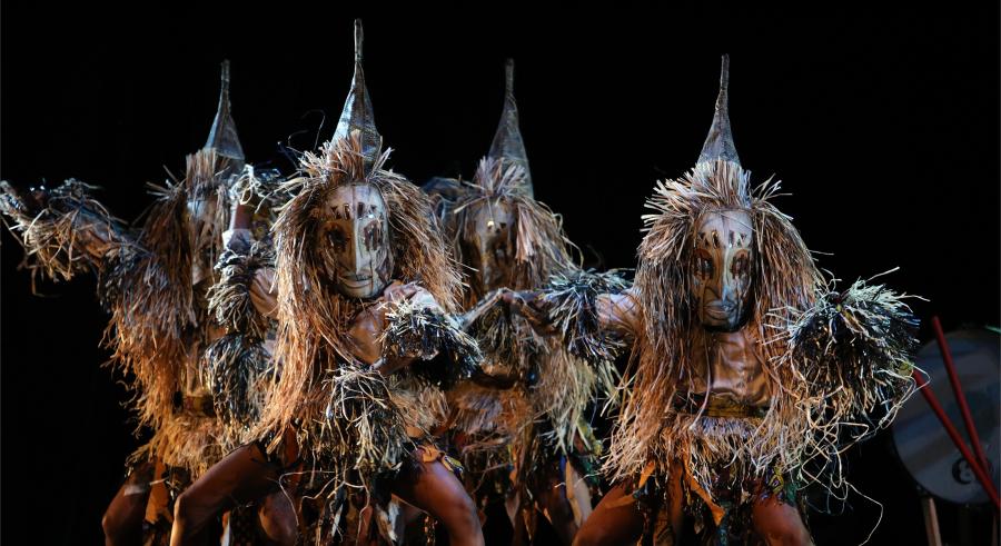 A group of dancers wearing creature-like costumes with pointed headwear appear onstage, facing the camera with their legs bent out toward their sides.