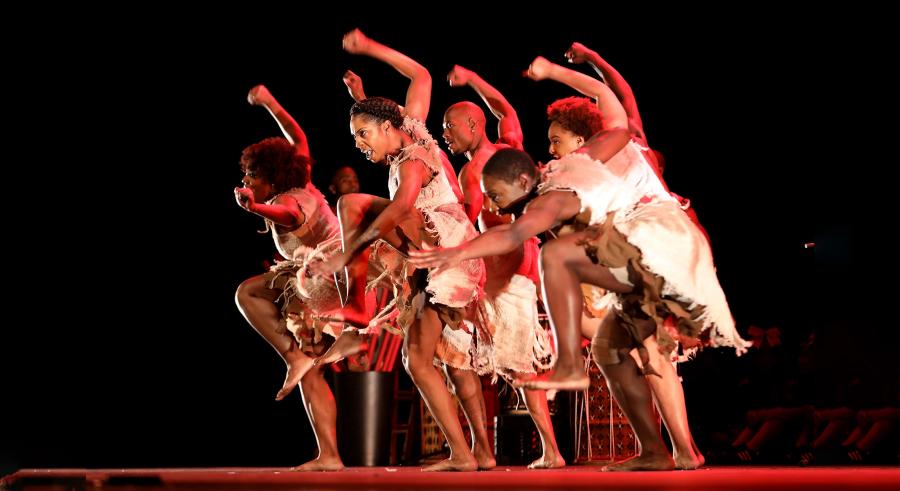 A group of dancers stand closely together onstage dressed in white clothing with their right arms and left legs raised, gazing downward toward the left and accompanied by a soft glow of red lighting.