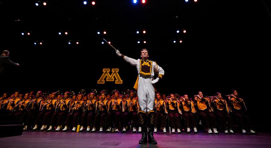 A drum major wearing white, maroon, and gold stands in a strong position with one arm lifted and the other bent towards their side. The marching band and the University of Minnesota letter "M" logo appears behind them.