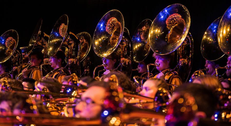 A close-up image shows marching band members wearing maroon and gold play their various brass horn instruments..