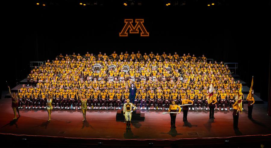 A marching band dressed in maroon and gold performs onstage with a University of Minnesota letter "M" logo above them. A drum major and a group of color guard members holding flags and batons stand in a line slightly downstage.