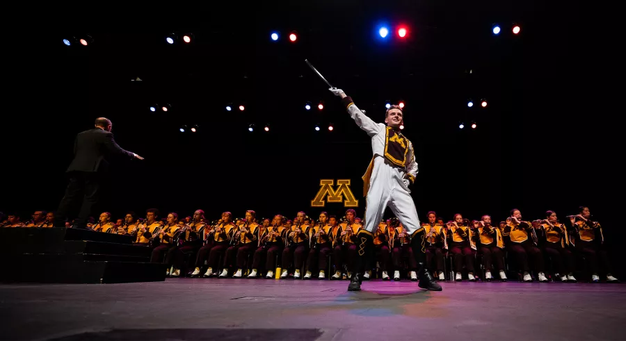 A drum major wearing white, maroon, and gold stands downstage with a large marching band grouped behind them. A University of Minnesota letter "M" logo appears in the background.