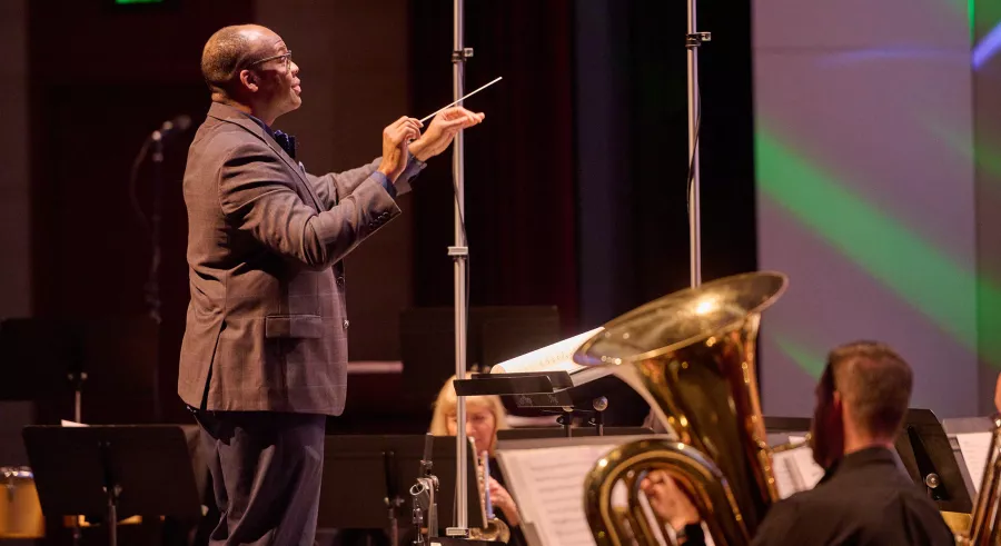 A person wearing a black suit faces to the right with their hands lifted and a conducting baton in one hand. A tuba player appears slightly below and to the right of the frame.