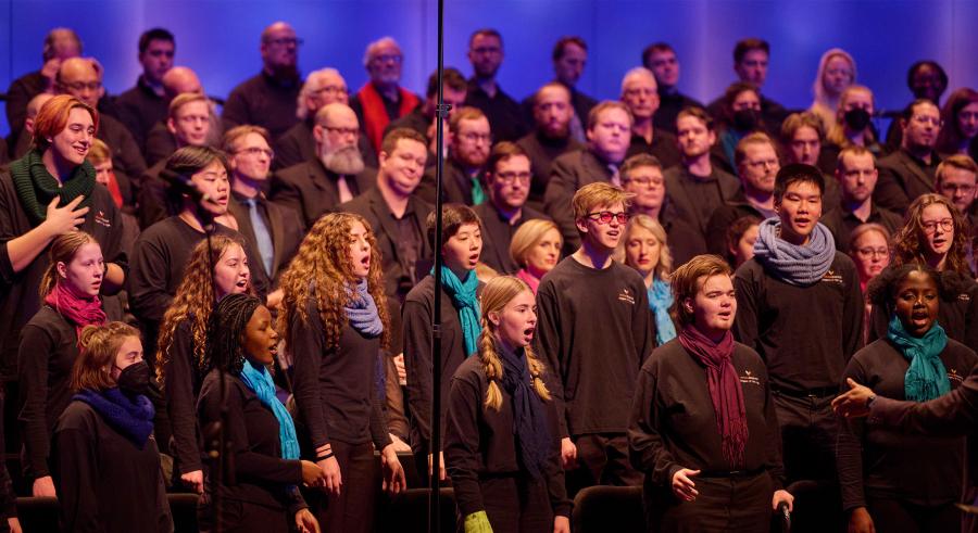 A group of chorus singers dressed in black with a mix of colored scarves perform with their hands resting to their sides, lined up in vertical rows of increasing height.