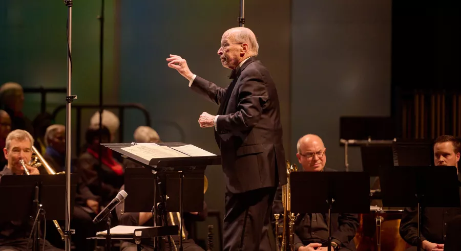 A conductor dressed in black faces to the left with one arm lifted and the other bent at their side. A music stand and musicians appear below them.