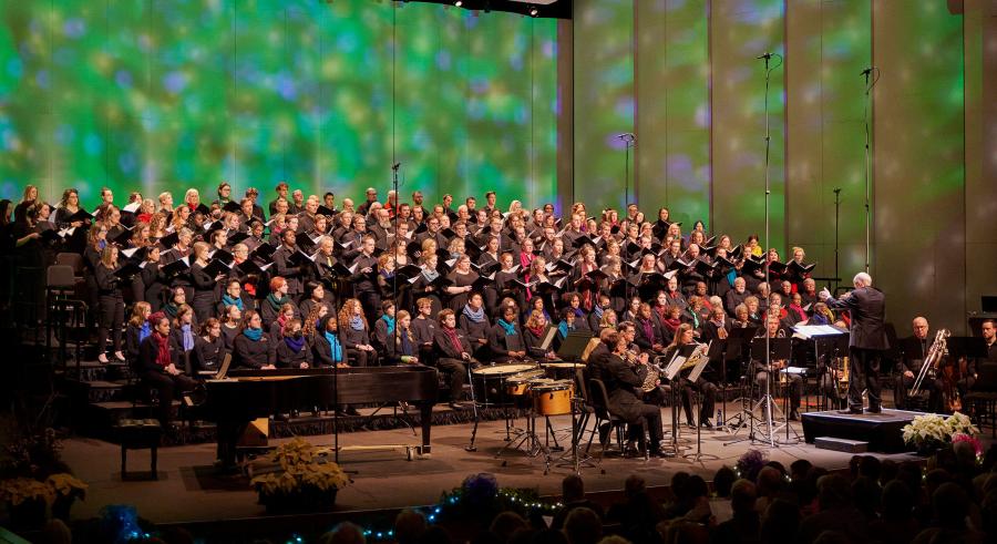 A choir wearing black with various colored scarves performs onstage with patterned green, blue, and yellow lighting projected behind them. A conductor stands downstage with their back toward the camera and musicians surrounding them. 