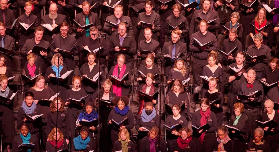 A group of chorus singers dressed in black with a mix of colored scarves perform with their music folders opened, lined up in verticle rows of increasing height. Pink and yellow projections appear behind them.