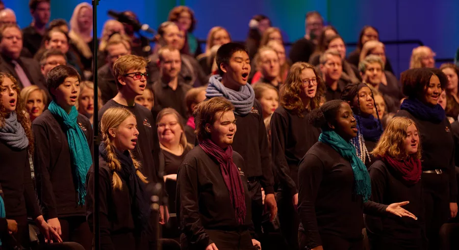 A group of chorus singers dressed in black with a mix of colored scarves perform with their hands resting to their sides, lined up in vertical rows of increasing height.