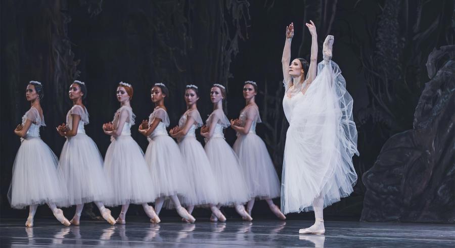 A ballerina extends her leg to the side above her head. She stands beside a line of identically posed dancers in white.