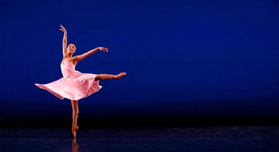 Solo female dancer in arabesque in a pink dress against a dark blue background.