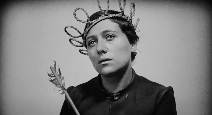 Black and white film still of Joan of Arc wearing a straw crown and holding an arrow.