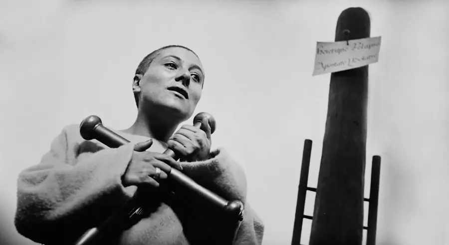 Black and white film still of a woman with shaved head wearing roughspun tunic holding a large cross in her arms in front of a tall post.