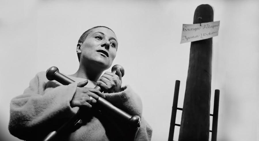 Black and white film still of a woman with shaved head wearing roughspun tunic holding a large cross in her arms in front of a tall post.