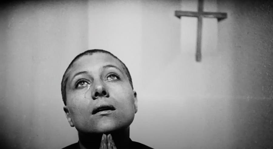 still from b/w film of woman with shaved head praying. A cross is on the wall behind her.
