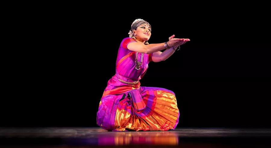 Dancer in purple kneels with both arms stretched forward on black stage.