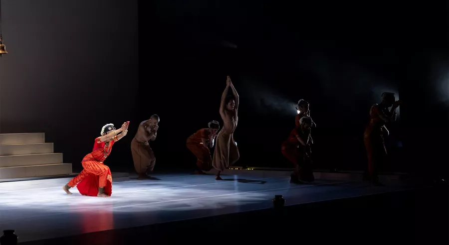 A dancer in red kneels in spotlight, others move in shadow.