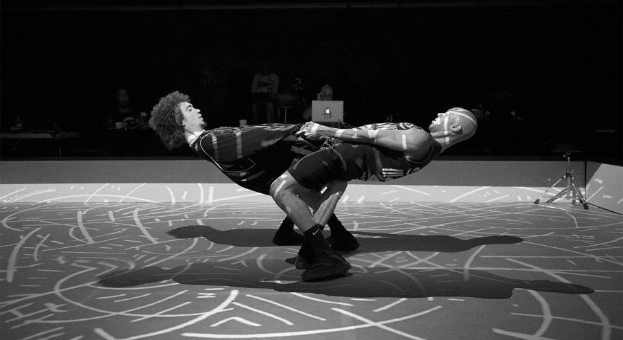 Black and white photo of dancers joining hands and leaning back on each other's weight, with lighted inscriptions projected onto their skin.