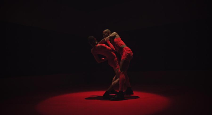 One dancer supports the other's frame. They are cast in half-dark red lighting.