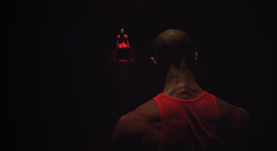Up-close picture of the back of a man's head, neck, and shoulders. He wears a red shirt and faces something glowing red in the dark.