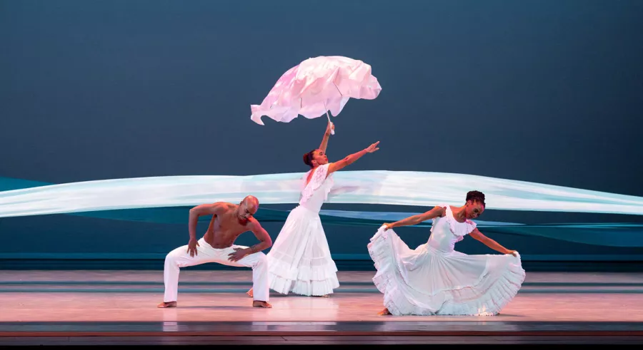 Dancer holding a pink umbrella with an arm reaching forward with a dancer on either side of her.